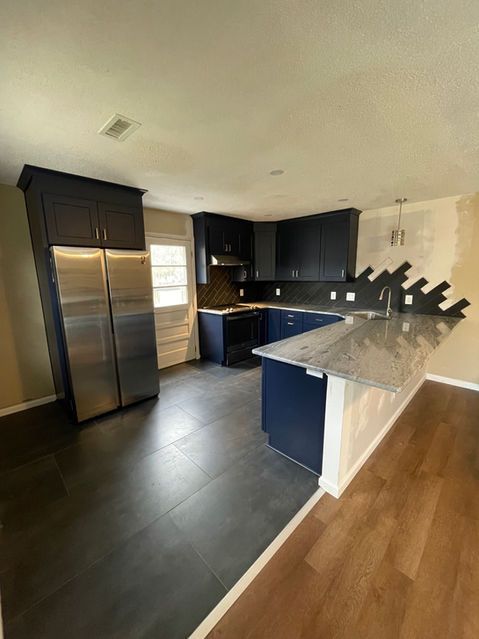 Empty modern kitchen with dark cabinets, stainless fridge, black counters, and light wood flooring