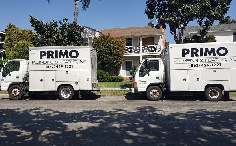Two white Primo Plumbing & Heating trucks parked on a street in front of a house.