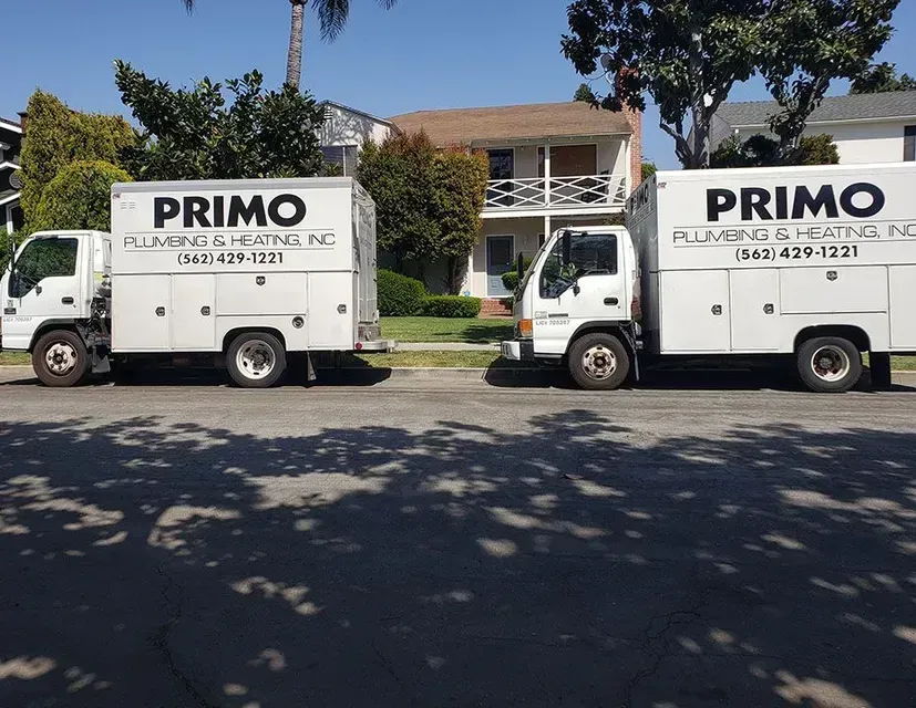 Two white Primo Plumbing & Heating trucks parked on a street in front of a house.