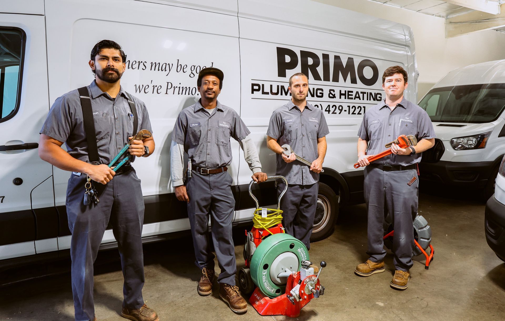 Four plumbers in gray uniforms standing with tools in front of a white Primo Plumbing and Heating van.
