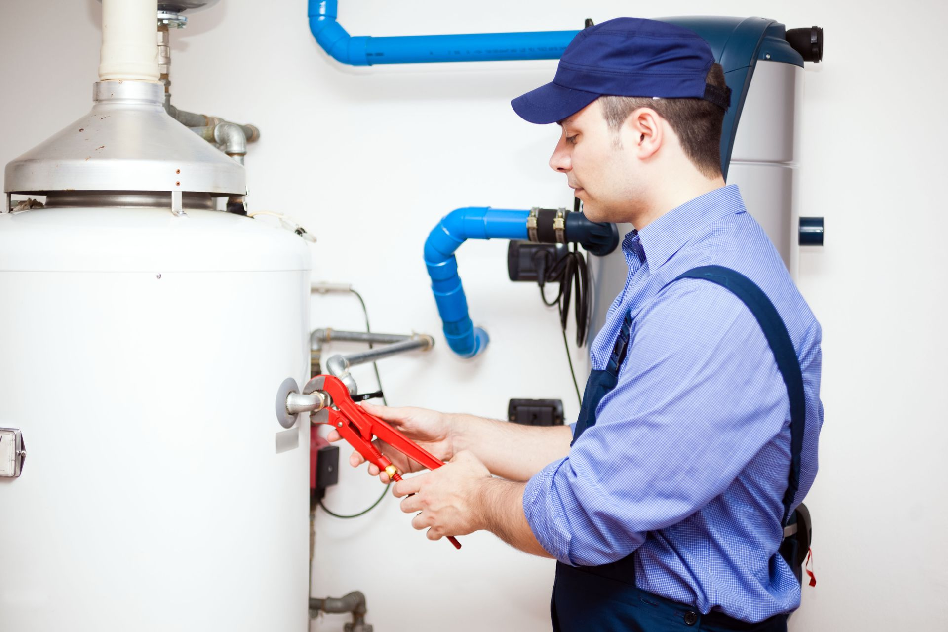 A professional in a blue uniform uses a red pipe wrench to work on a large, white water heater in a mechanical room.