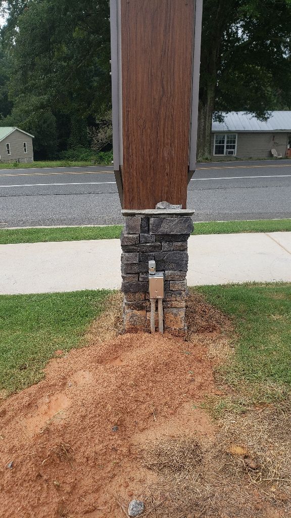 Stone and wood pillar with electrical box, in front of a yard and street.