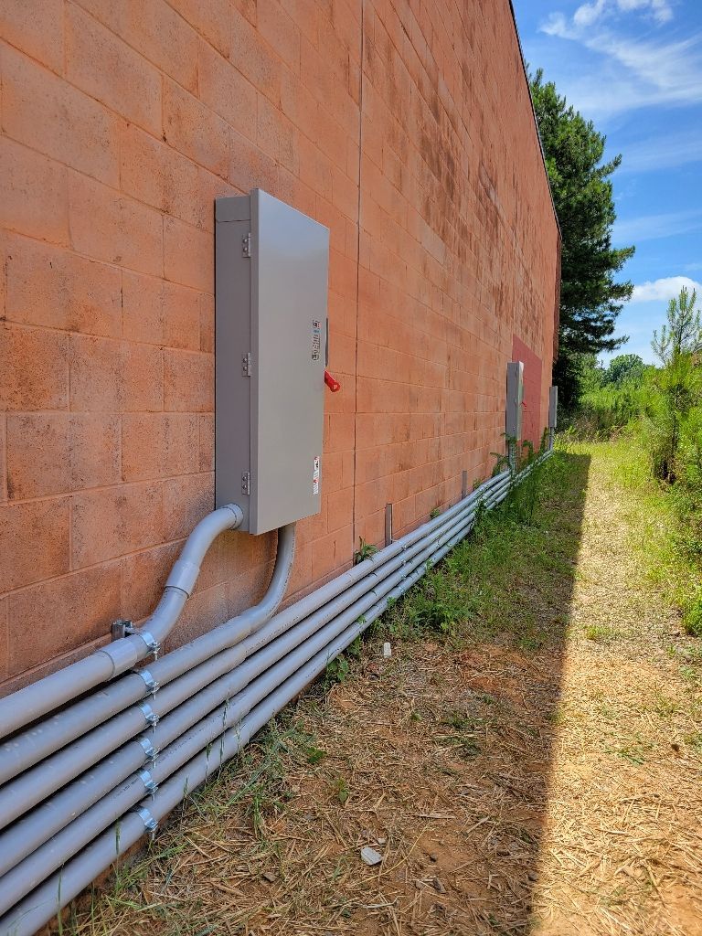 Gray electrical box and conduits on a brick wall beside a grassy path under a blue sky.