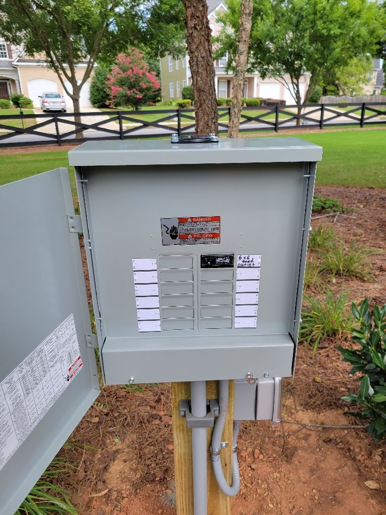 Gray electrical panel, door open, mounted on wooden post in a yard.