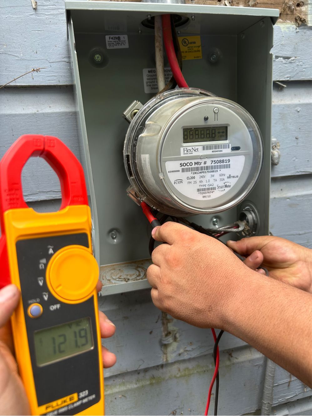 Electrician using a clamp meter to measure current on a home's electrical meter in an outdoor setting.
