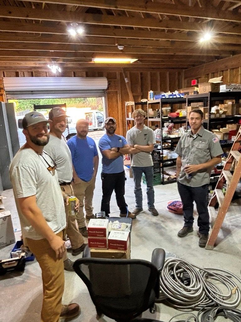 Group of men in a garage setting, some in work attire, looking at a table with equipment.