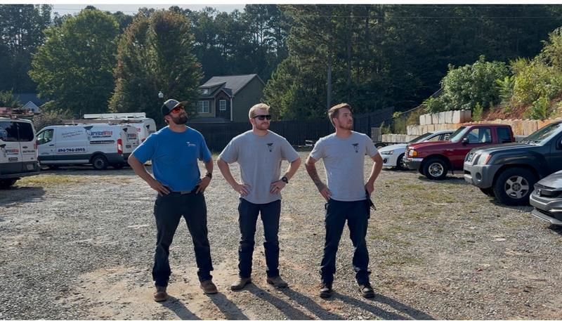Three men standing in a parking lot, hands on hips, looking up, with vehicles and a building in the background.
