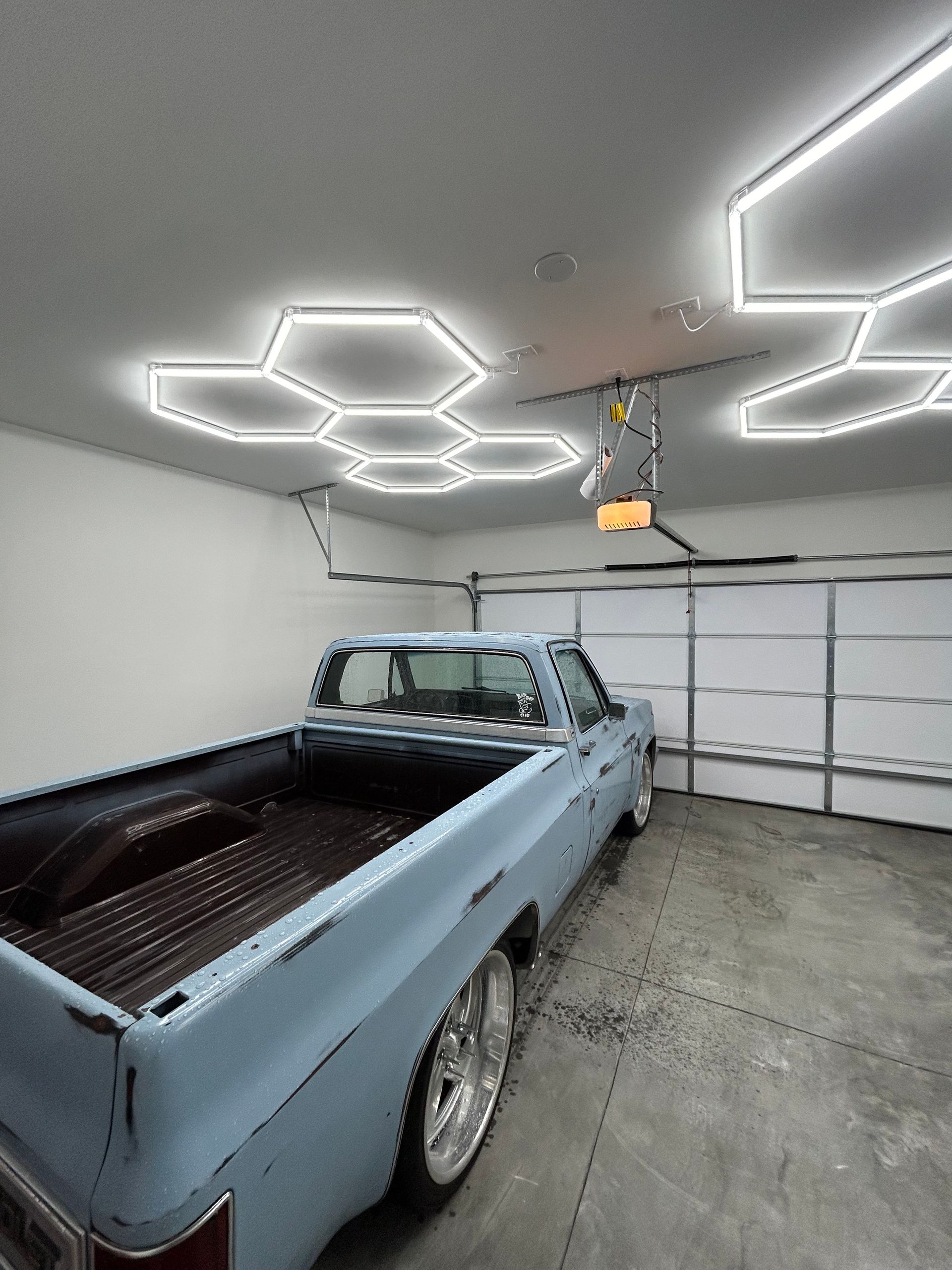 Light blue pickup truck inside a garage with hexagon-shaped ceiling lights. Garage door is open.