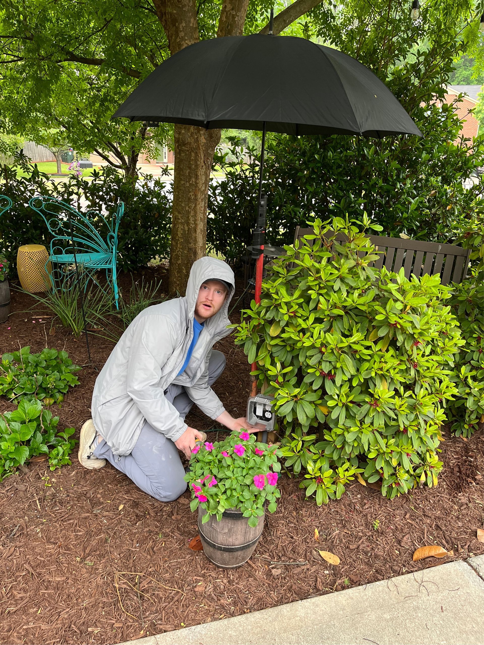 Person kneeling, tending flowers. Umbrella over a plant near a tree.  Green foliage and a blue bench in the background.