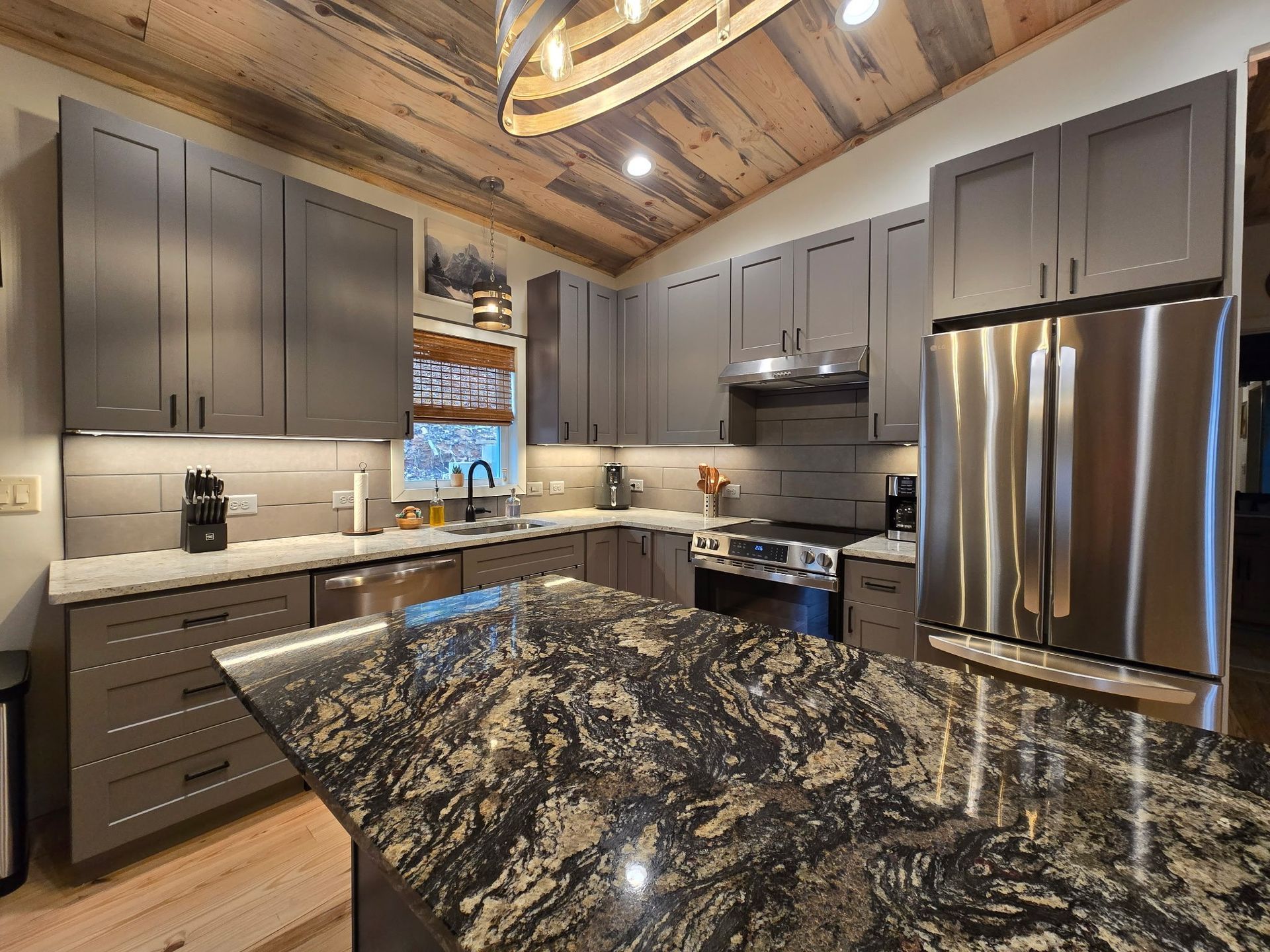 Modern kitchen with gray cabinets, granite island, stainless steel appliances, and wood ceiling.