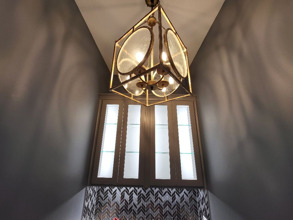 Ornate geometric chandelier above tall, narrow windows with frosted glass. Dark walls, patterned curtain.