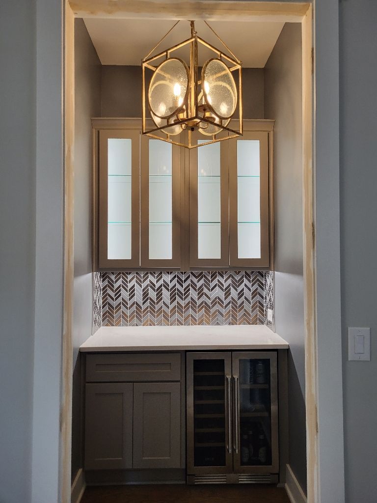 Built-in bar with gray cabinets, a wine fridge, and a decorative tile backsplash under a gold chandelier.
