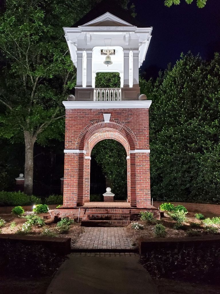 Brick bell tower at night, lit from below, with archway entrance and white-painted top.