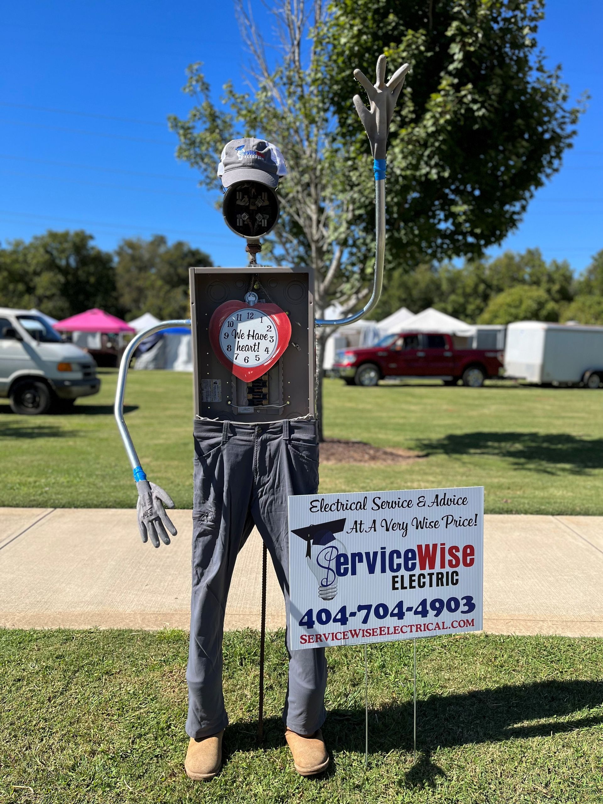 Scarecrow-like figure made of electrical parts holds ServiceWise Electric sign on grass.