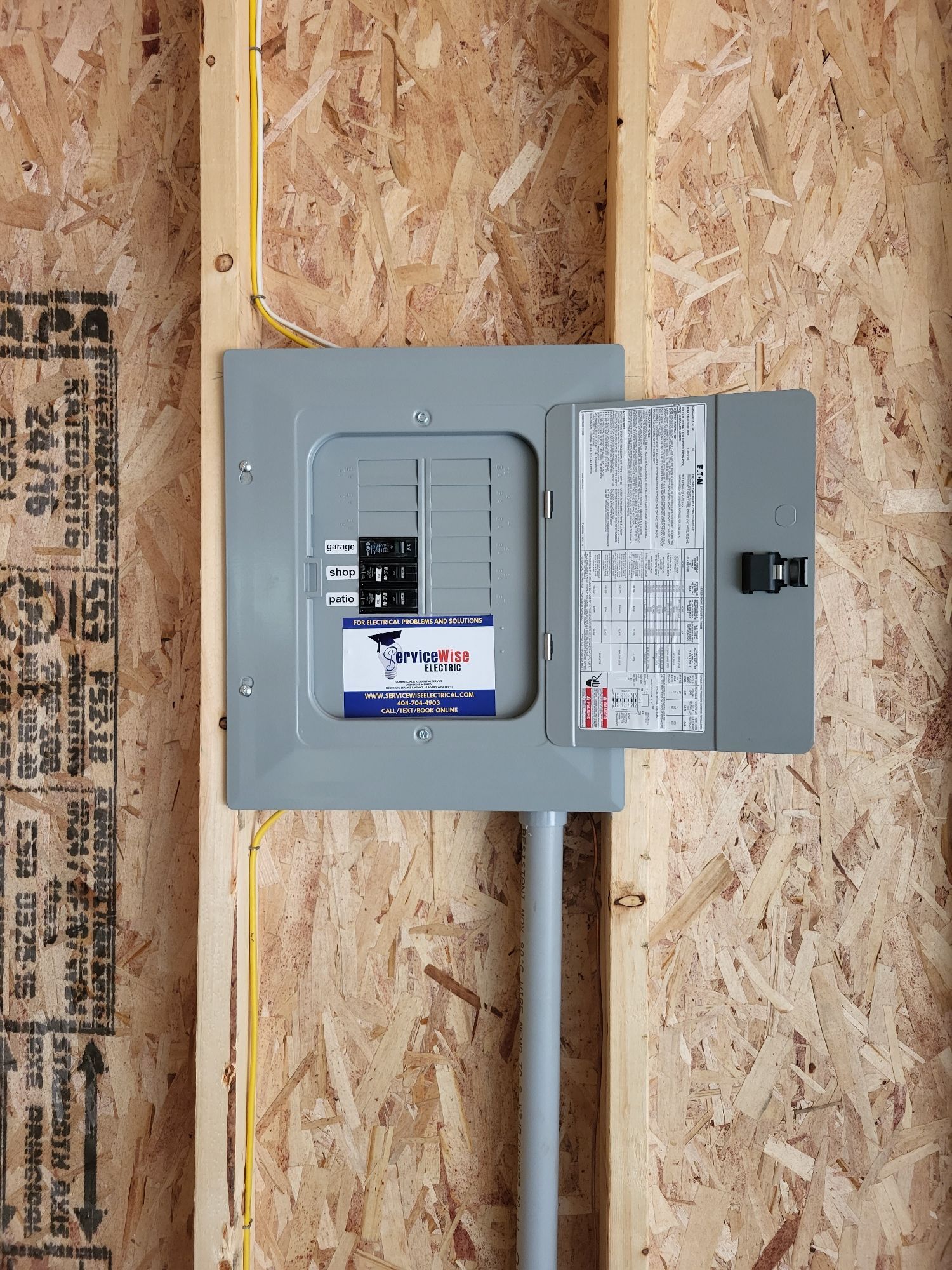 Electrical panel installed in a wall; gray metal box, open door with circuit breakers.