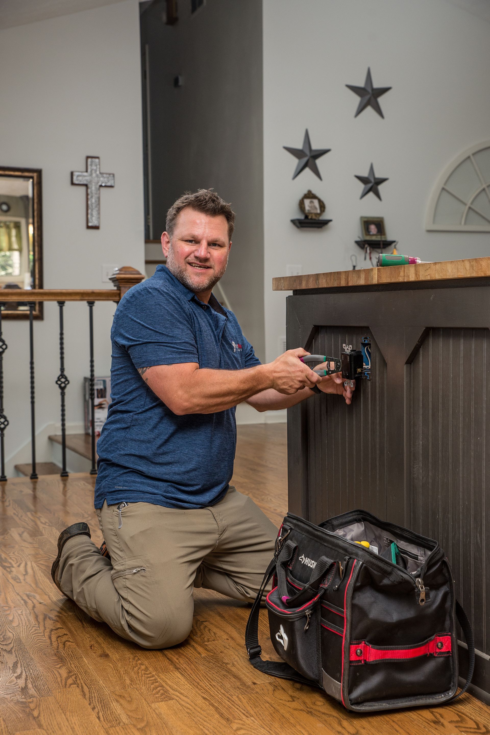 Man kneeling, working on outlet in a kitchen. Tool bag beside him, wooden floor.