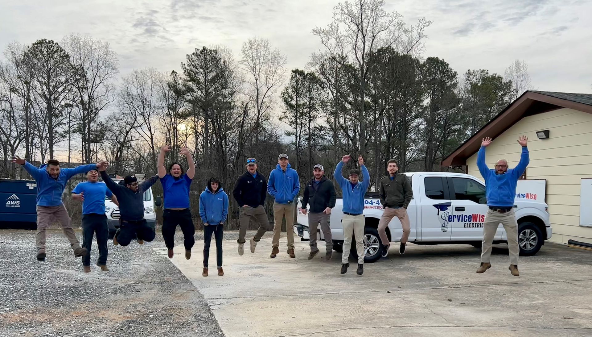 Group of people in blue shirts jumping with arms raised, near a white truck, trees, and a building.