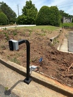 Black curved metal post with attached gray boxes; next to a curb and dirt landscaping.