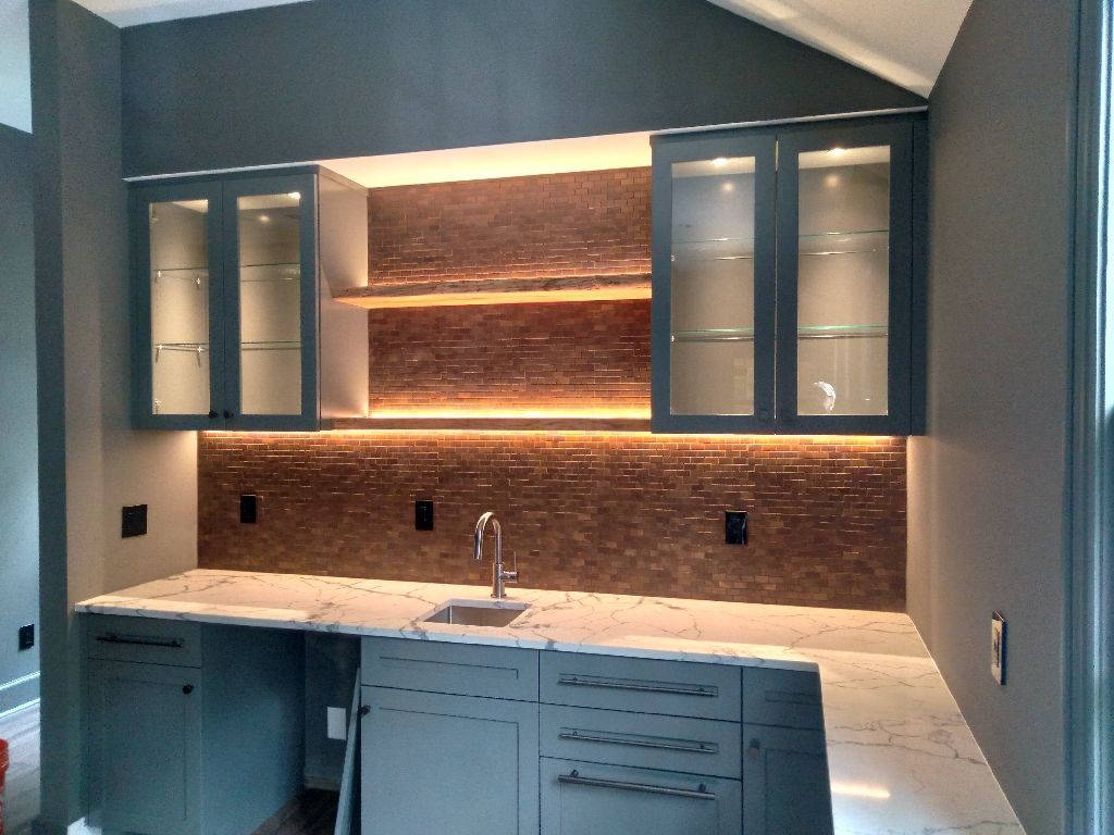 Gray built-in bar with upper glass-door cabinets, shelves, brown tiled backsplash, and sink on white countertop.