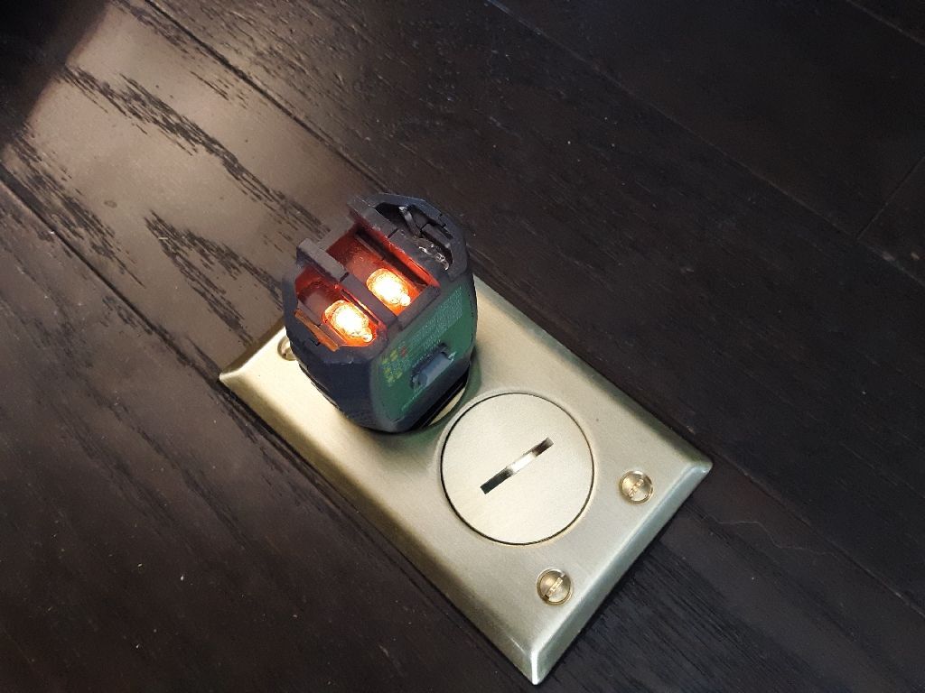 A floor outlet with a device plugged in and two orange lights illuminated on dark wooden floor.