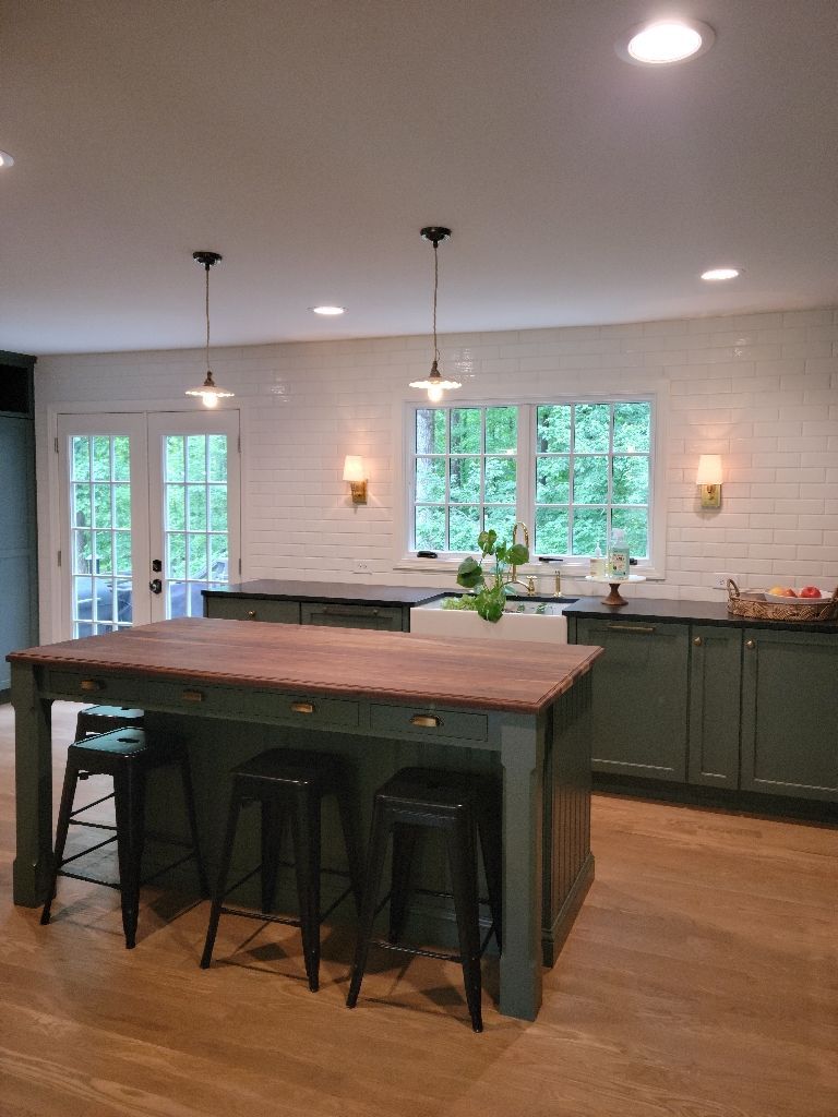 Green kitchen with wooden island, black stools, white backsplash, and windows overlooking greenery.