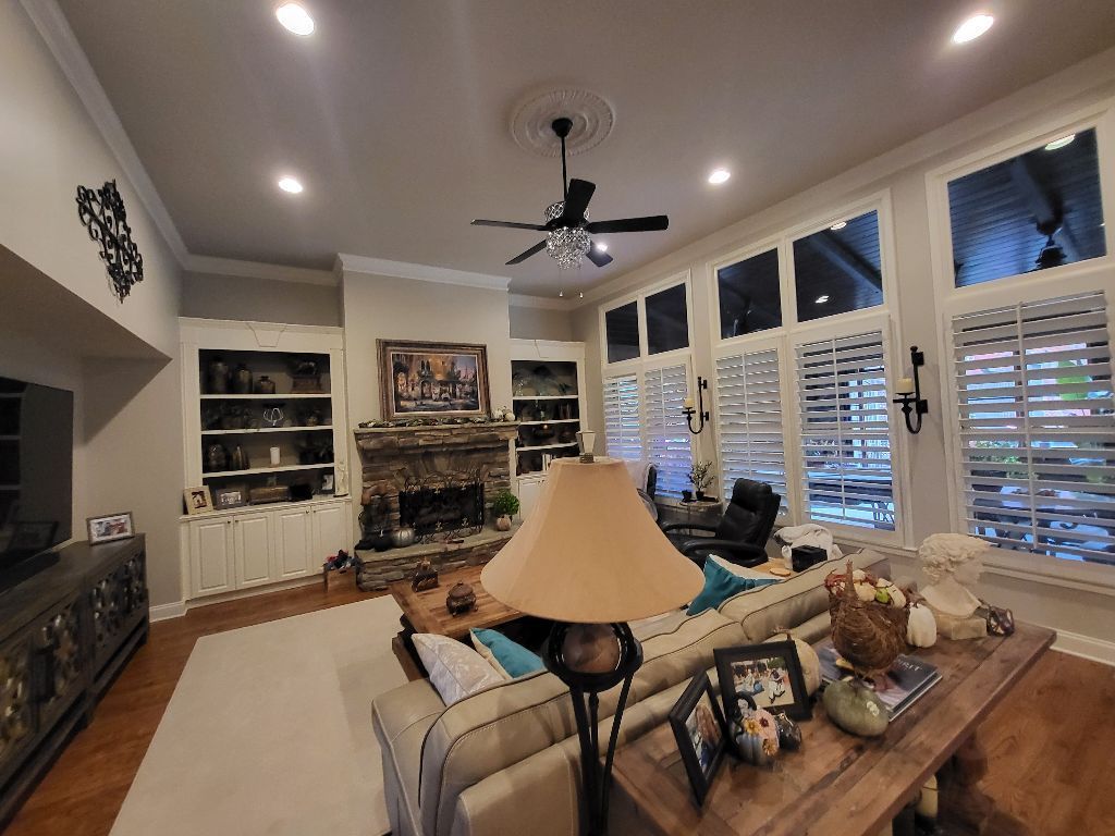 Living room with fireplace, built-in shelves, sofa, and large windows with shutters; warm lighting.