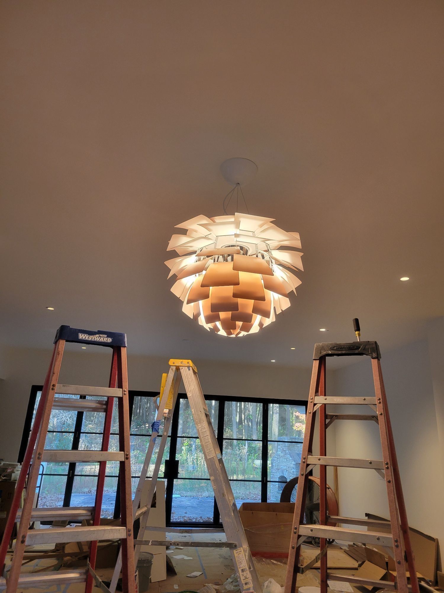 Three ladders surround a modern chandelier on a white ceiling. Large windows in the background.