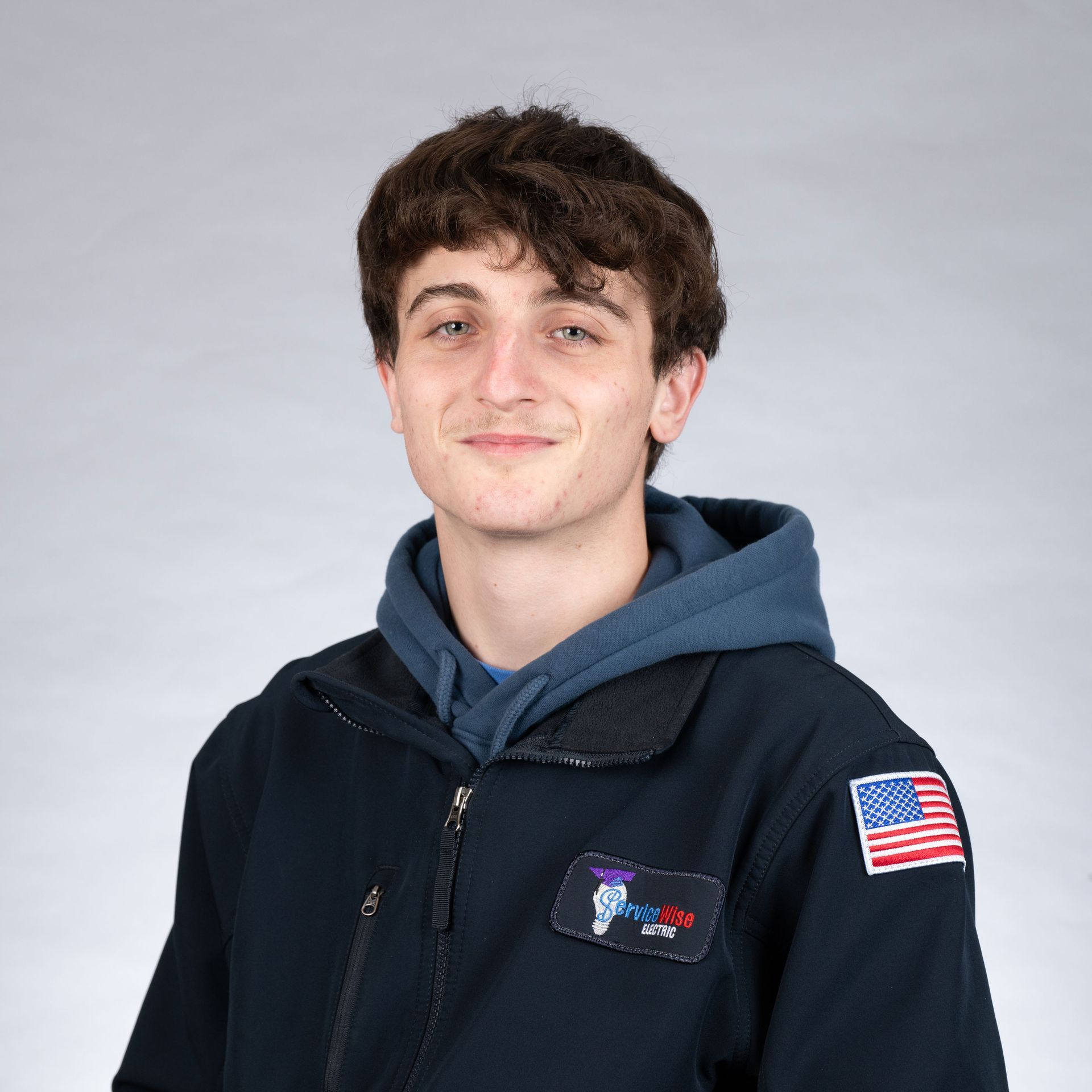 Young person with curly brown hair, smiling, wearing a blue hoodie and navy jacket with an American flag patch.