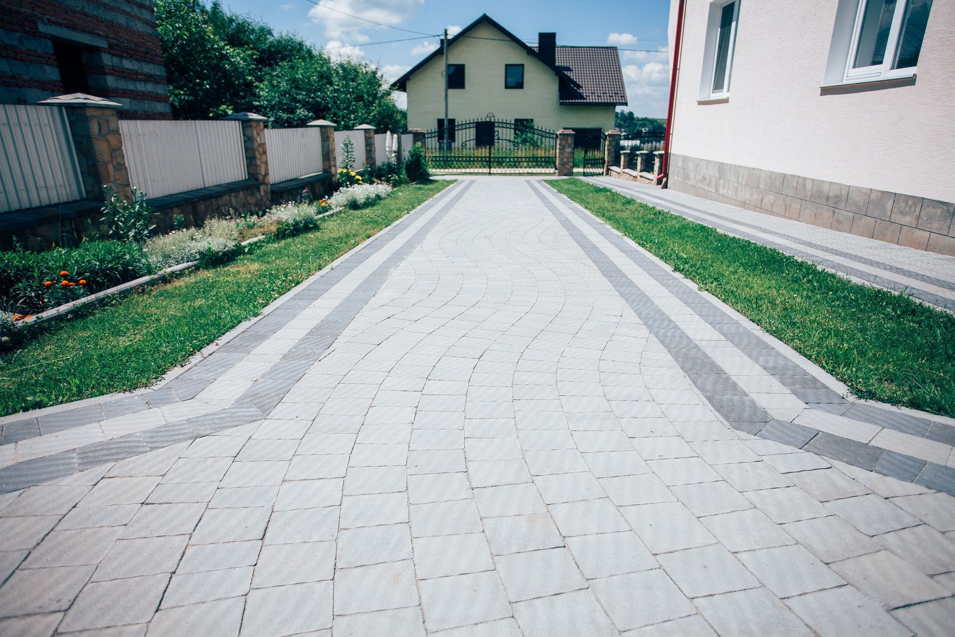 Paved driveway with gray stone leading to a yellow house, bordered by grass and a low fence.