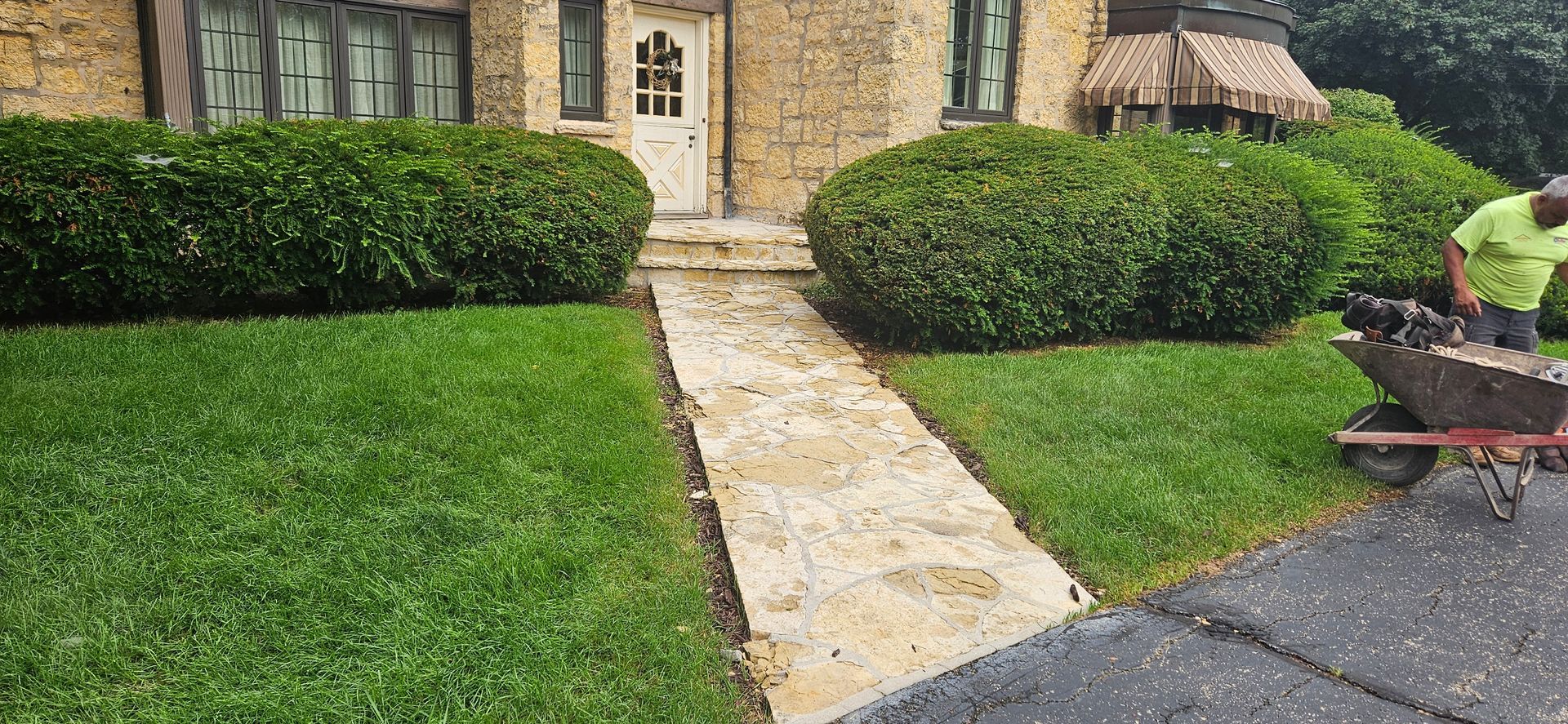 A man is pushing a wheelbarrow down a sidewalk in front of a house.