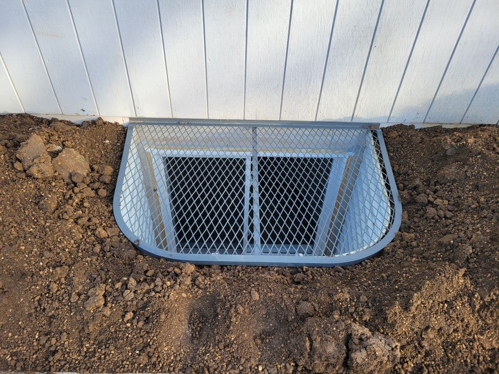 A window well is sitting in the dirt next to a white fence.