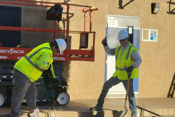 Two construction workers by a lift, one holding a tool and the other striking a pose with a thumbs up near a building entrance.