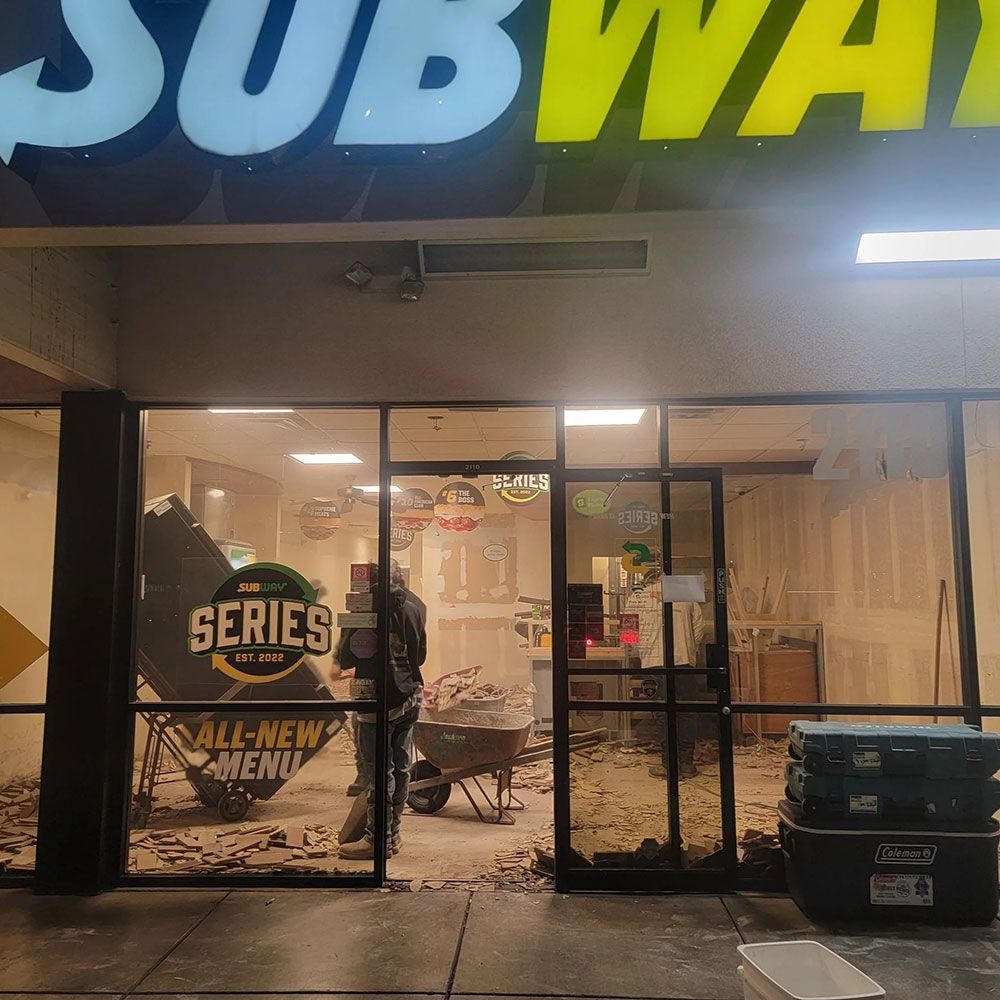 Subway storefront damaged, worker inside clearing rubble. Exterior shot, sign visible.
