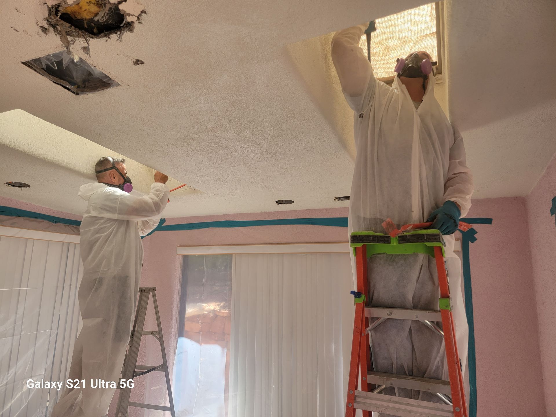 Two people in protective suits removing ceiling tiles; interior, one on a ladder.