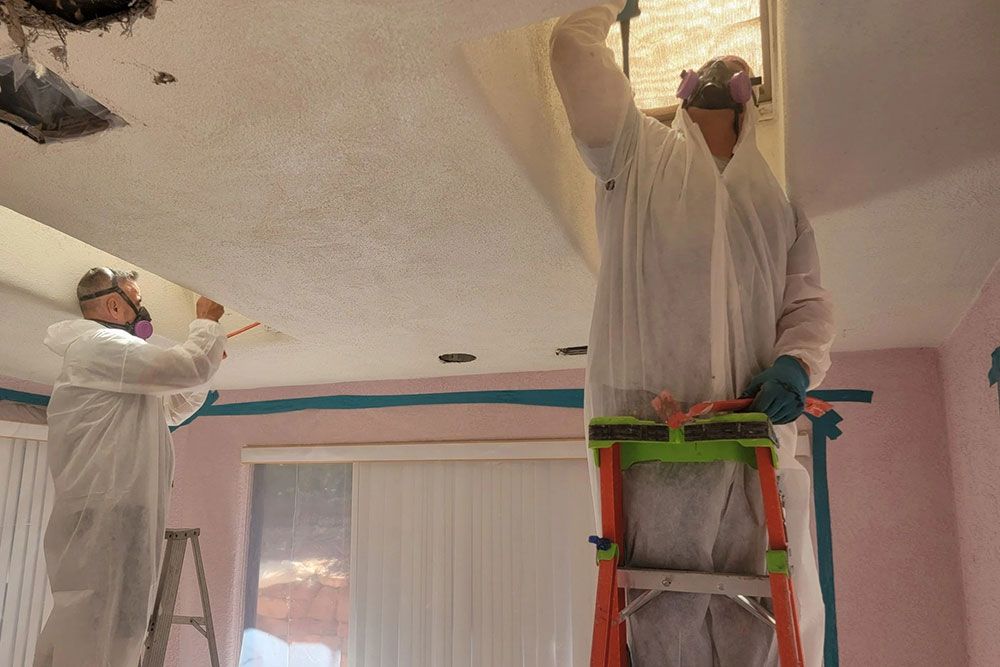 Two people in protective suits remove ceiling panel in a room. One stands on a ladder. Blue tape protects the walls.