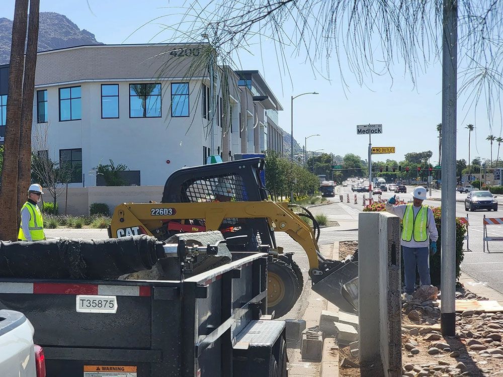 Construction workers and a yellow CAT skid steer loading debris into a trailer on a city street.