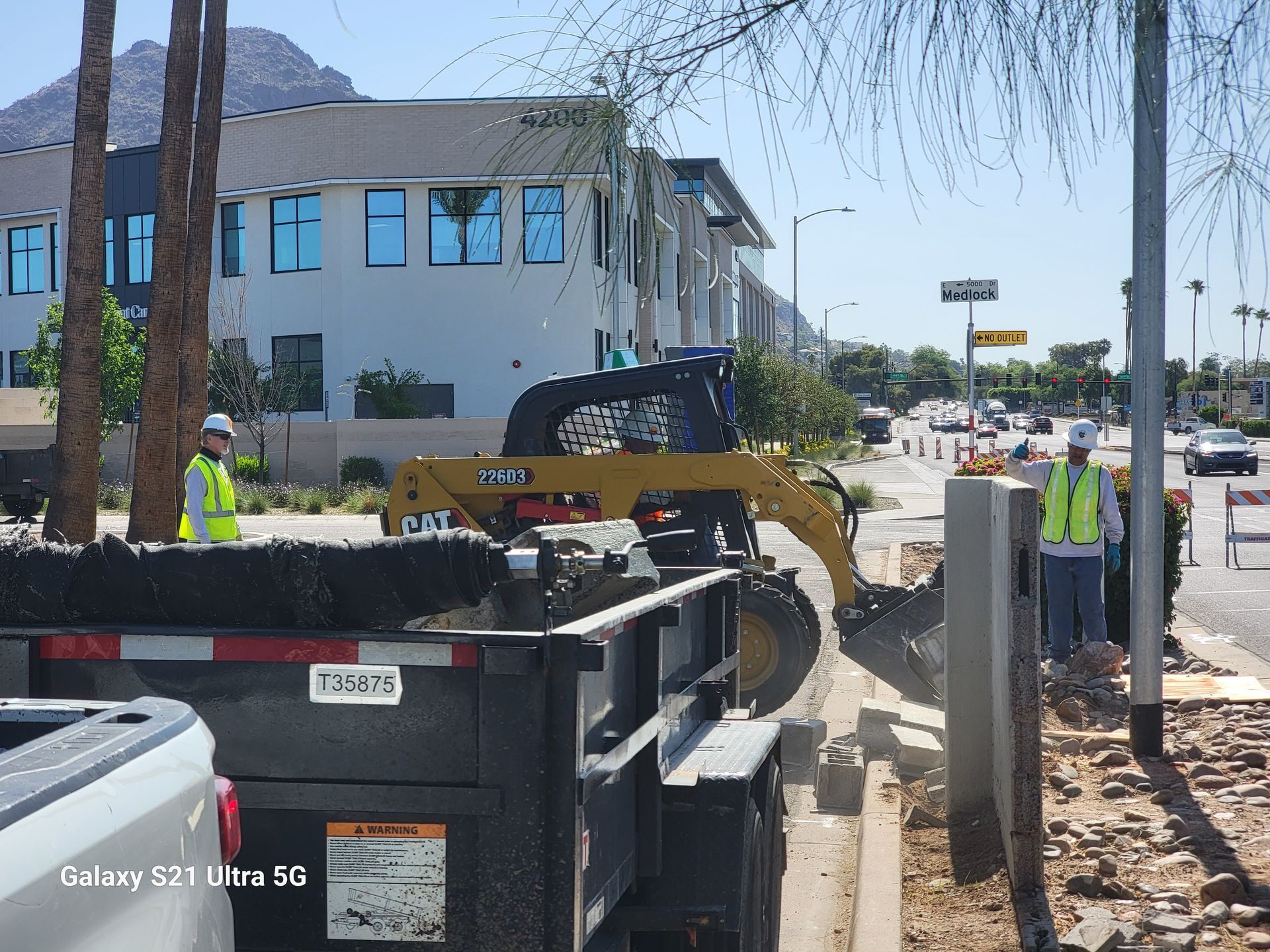 Construction site with a skid steer, workers, and a trailer on a street next to a building.