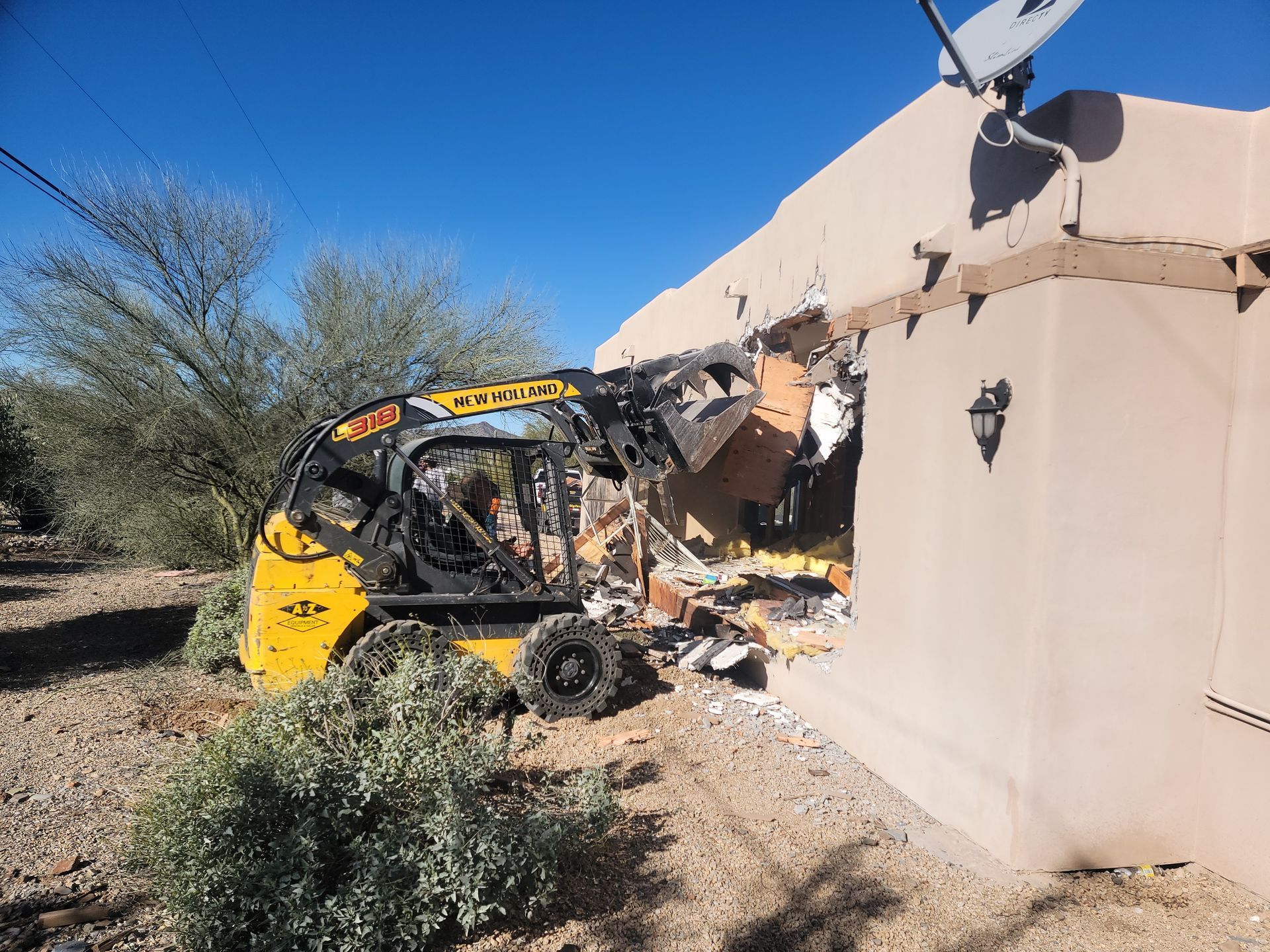 Yellow skid steer crashed into a tan building, creating a hole in the wall. Outdoors, sunny.