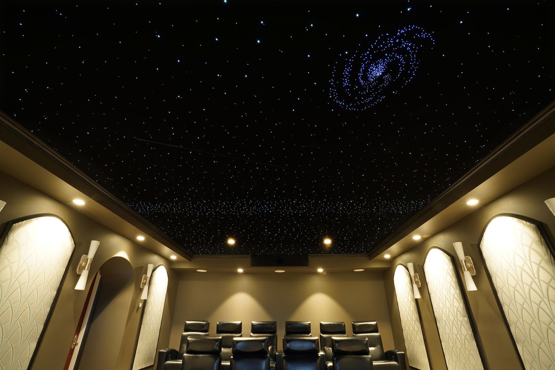 Home theater room with starry ceiling and galaxy design. Rows of black seats, beige walls.