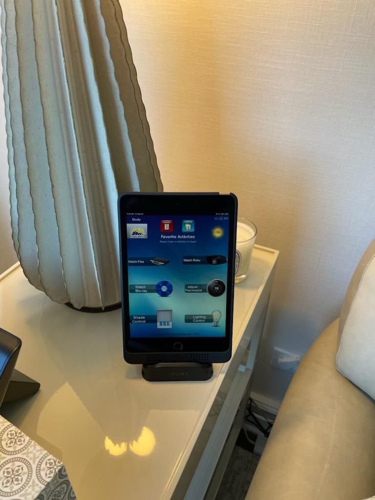 Tablet in black dock on a white table, showing home screen apps. Next to a cream lamp and a candle.