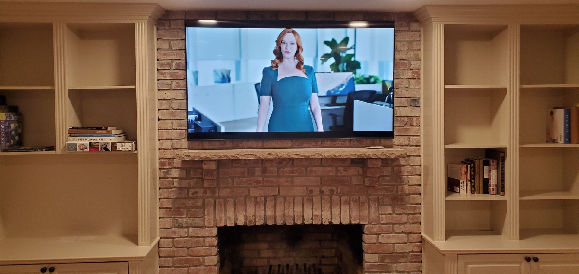 A TV above a brick fireplace with built-in shelves on either side. A woman is visible on the TV screen.