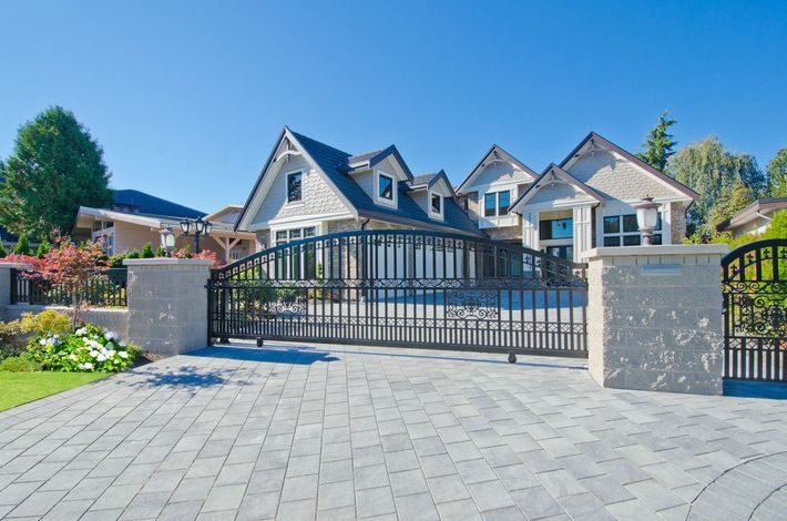 Large, gated house with gray brick driveway under a blue sky.