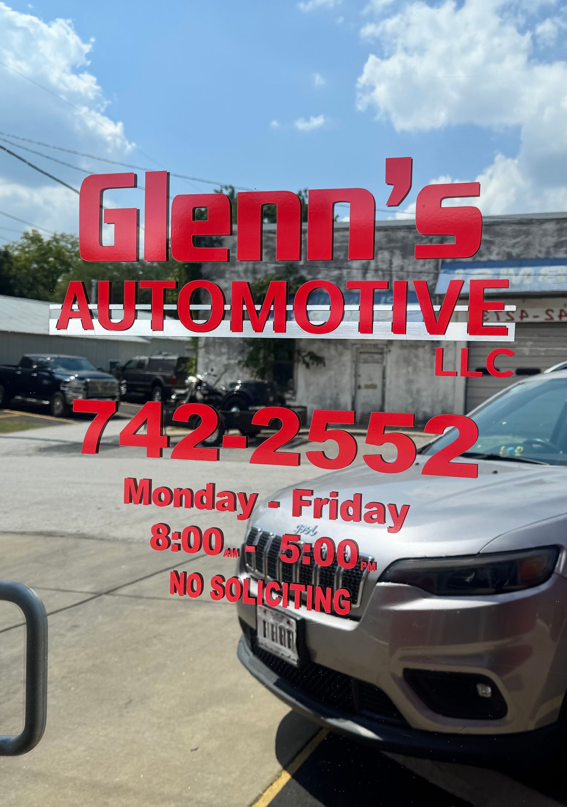 Glenn’s Automotive LLC storefront sign with phone number and hours, reflected in a glass window.