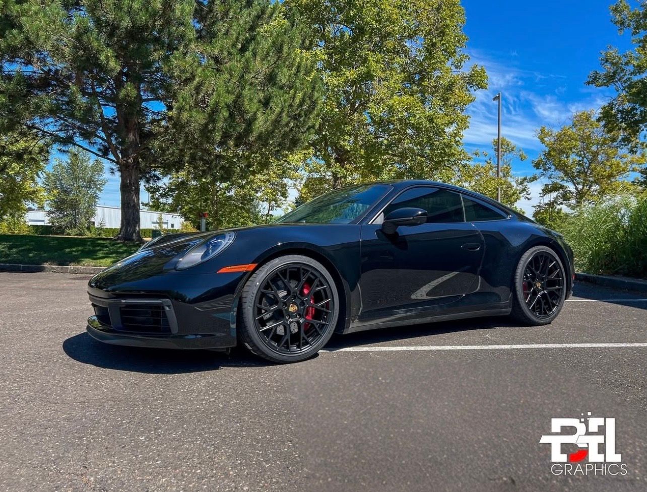 Black Porsche 911 parked on asphalt, red brake calipers visible, blue sky background.