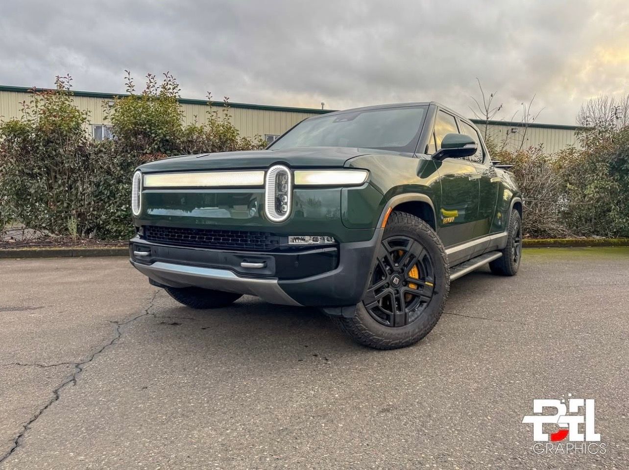 Green Rivian electric truck parked on asphalt. Black wheels, yellow calipers, cloudy sky.