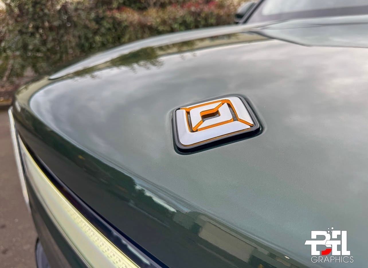Close-up of a Rivian truck hood with the logo in white and orange. Green paint is reflecting the sky.