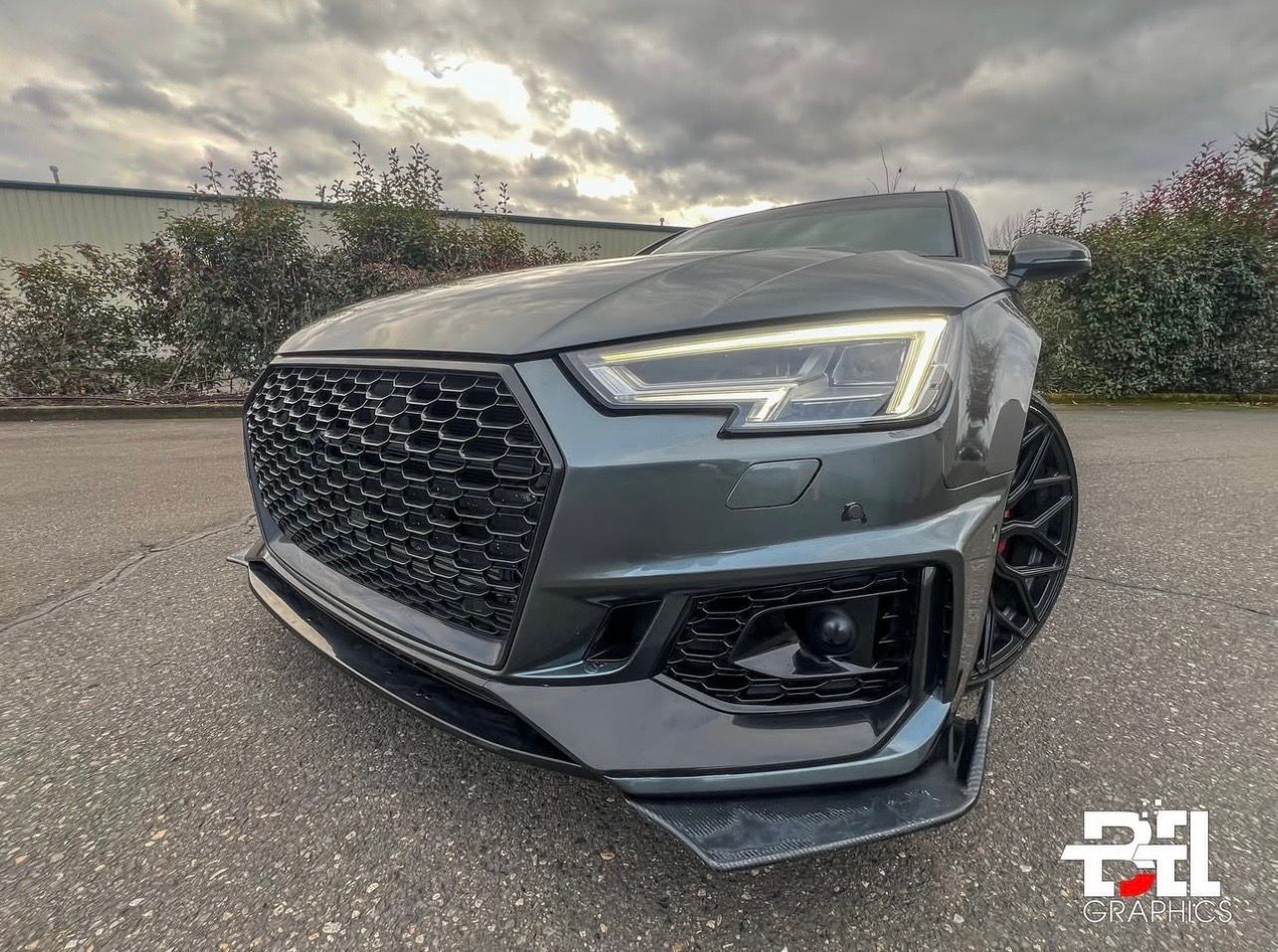 Gray Audi RS4 with black grille, front spoiler, and black wheels, parked outdoors under overcast skies.