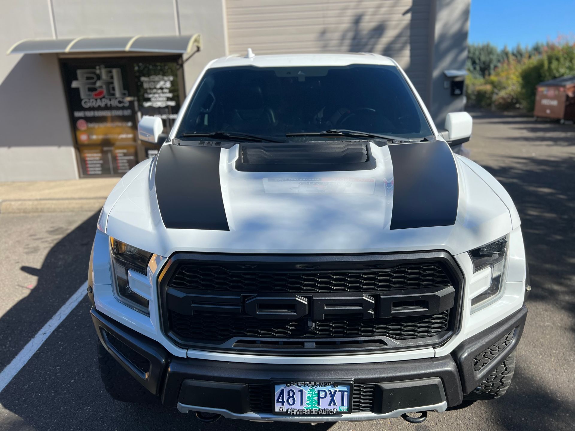 White Ford Raptor truck with black hood stripes and grille; parked in front of a building.