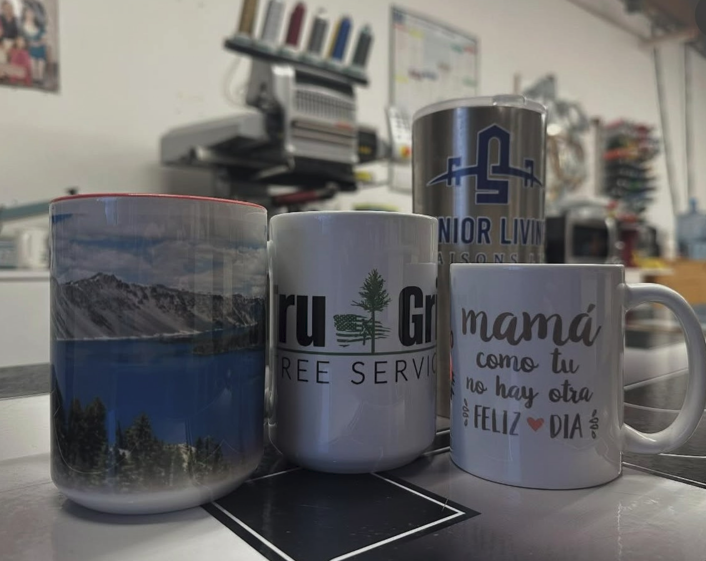 Four custom mugs with various designs in front of an embroidery machine.