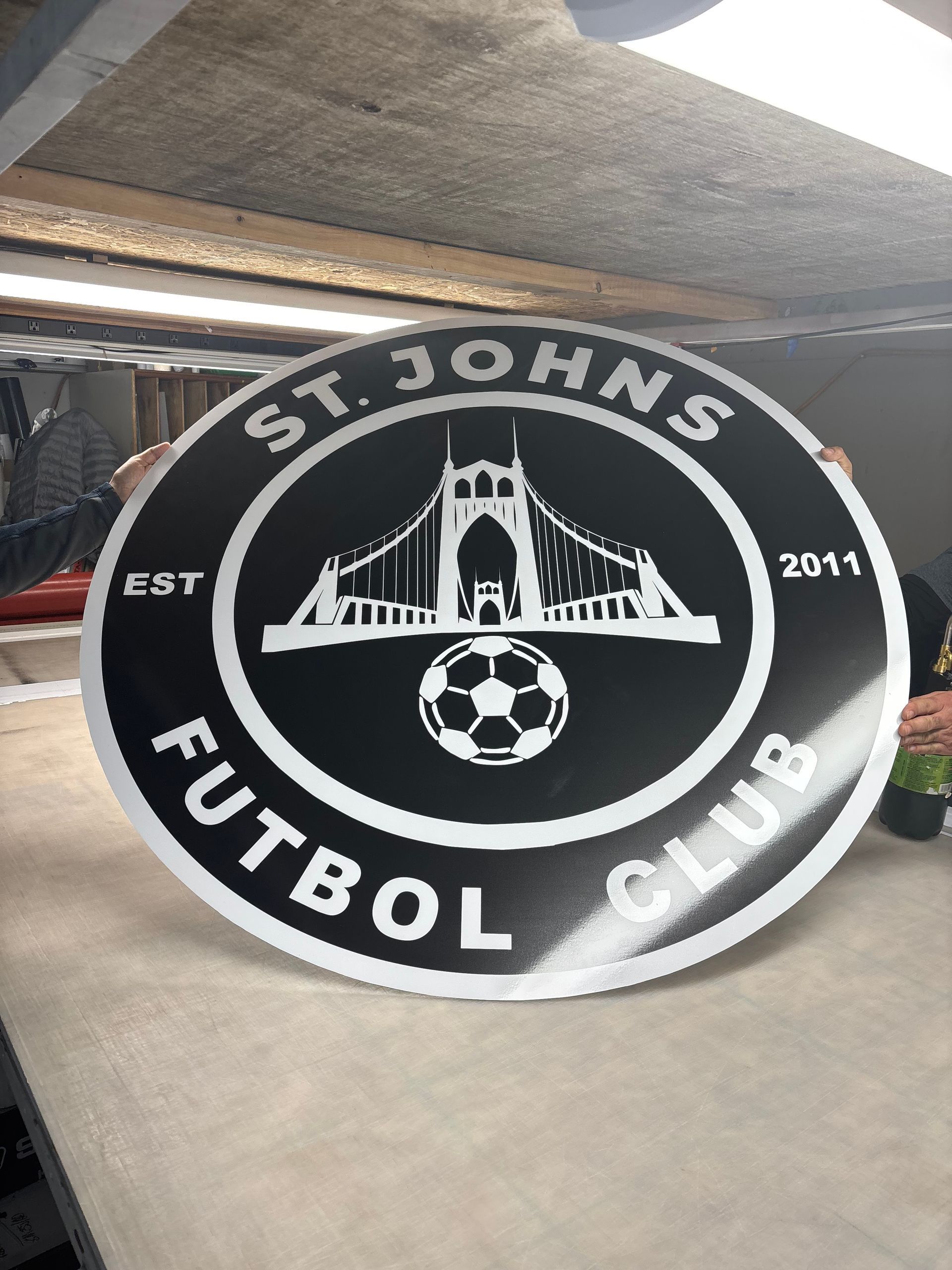 Round black and white sign for St. Johns Futbol Club. Features a bridge, soccer ball, and text.